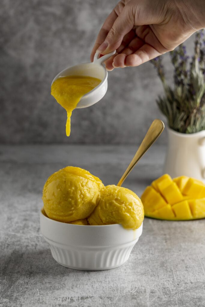 A bowl of creamy homemade mango ice cream served with fresh mango slices, with mango puree being drizzled over the ice cream.