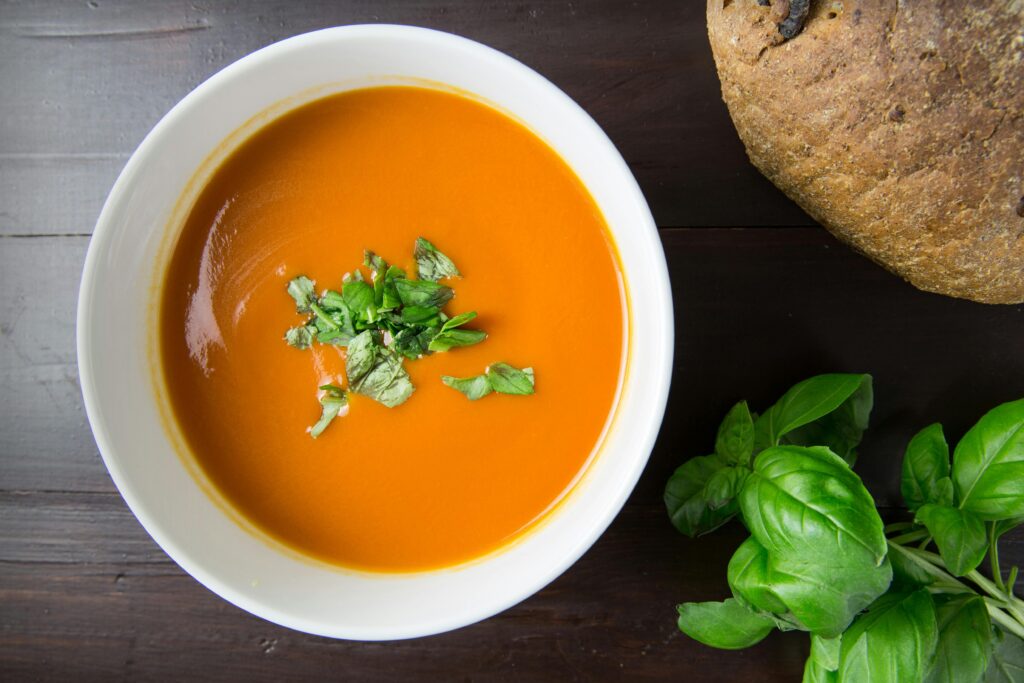 A bowl of creamy pumpkin soup garnished with roasted pumpkin seeds and fresh herbs, surrounded by fresh carrots, potatoes, and pumpkin slices.